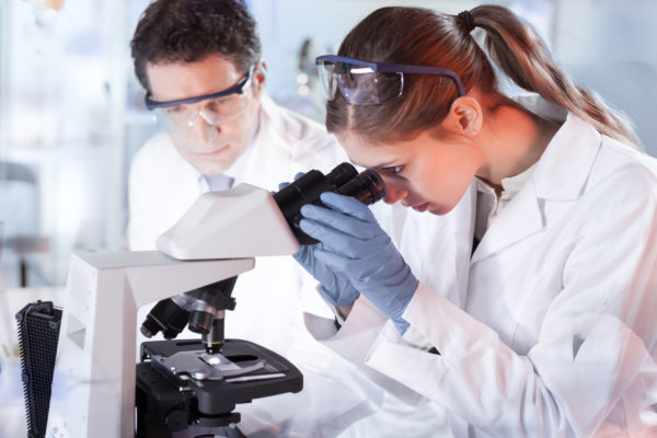 Life scientists researching in laboratory. Attractive female young scientist and her post doctoral supervisor microscoping in their working environment. Healthcare and biotechnology.